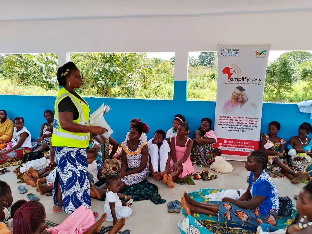 Séance de sensibilisation communautaire de MCF-CI : une agente de santé en gilet anime un groupe de femmes assises devant un centre de santé en Côte d'Ivoire